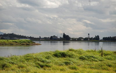 Imagen de un paisaje con una ladera son pastos al lado de una río y al fondo se visualizan casa y construcciones.